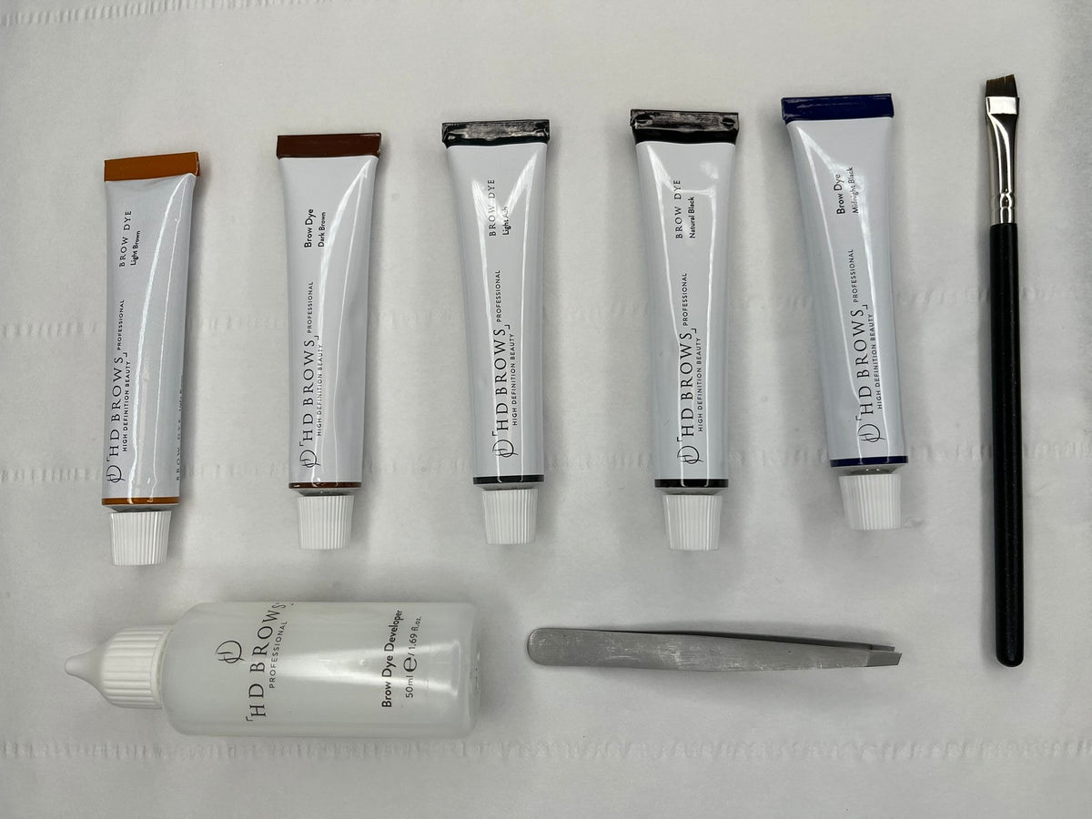 A brow grooming kit by HD BROWS arranged on a white textured background, including a clear bottle of brow dye developer, five white tubes of brow pigment with brown caps, a metal spatula, and two small applicator brushes.