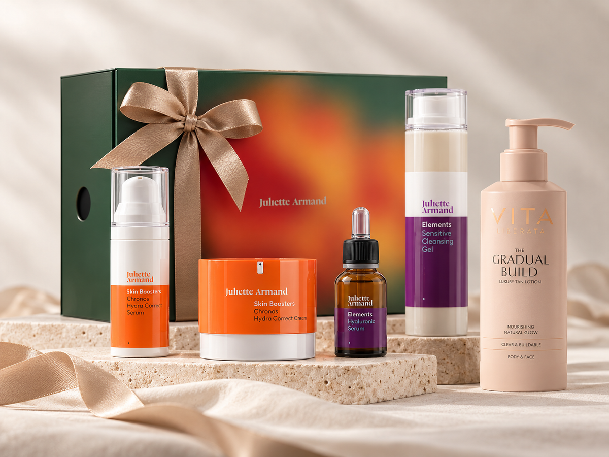 A curated skincare gift set from Juliette Armand, featuring a dark green gift box with a gold ribbon, a white pump bottle with orange accents, an orange box, a purple dropper bottle, a tall white bottle with purple labeling, and a large beige pump bottle labeled 'VITA The Gradual Build', all arranged on a stone slab with draped satin fabric.