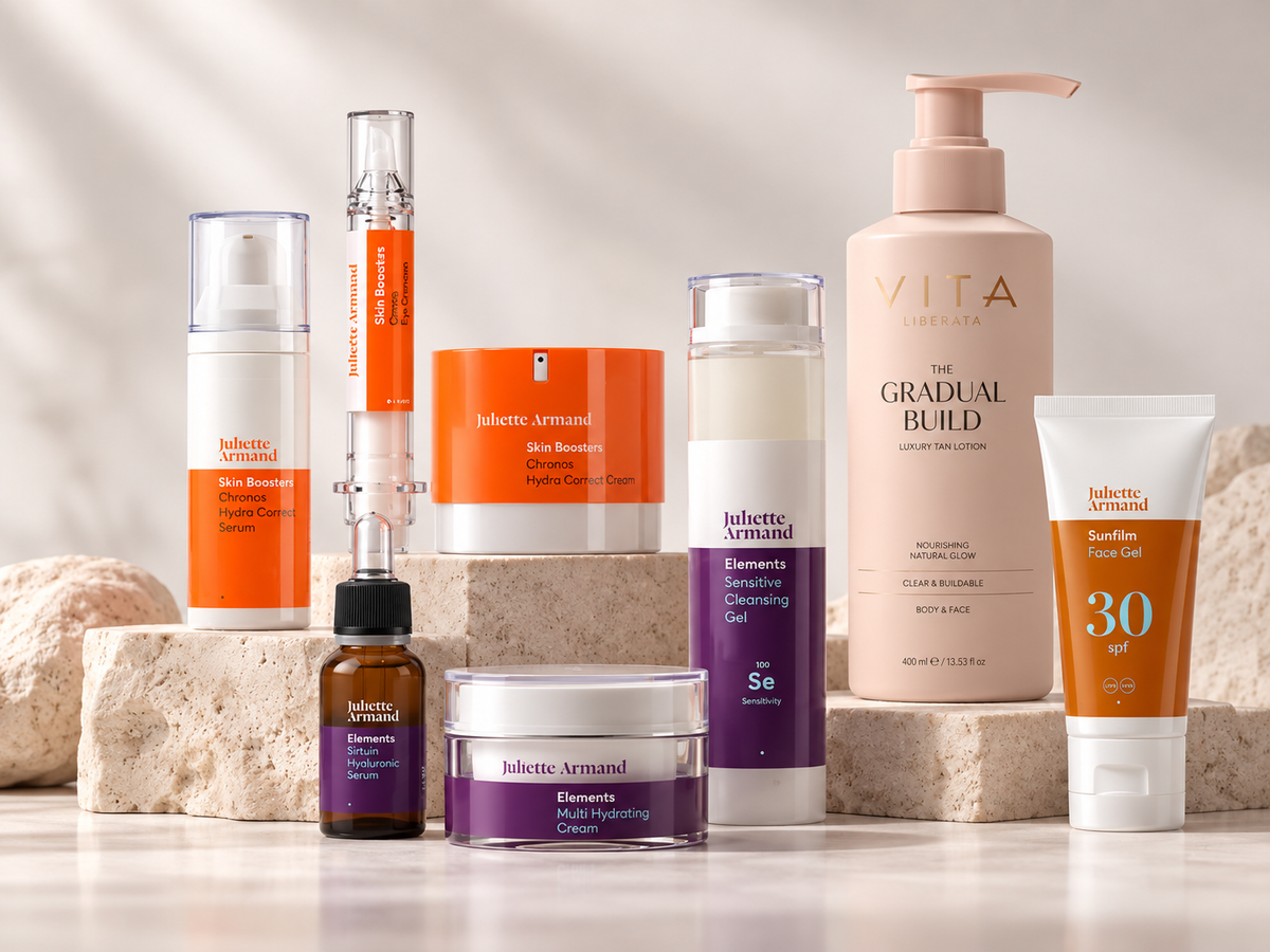 A curated collection of Jolette Armand and VITA skincare products arranged on light stone blocks against a warm, sunlit neutral background. The products include an orange and white Skin Booster pump bottle, an orange and white Gradual Build moisturizer pump bottle, a purple and white Se Accelerating Dermatology Gel tube, a purple and white Multi-Faceting Cream jar, a small amber bottle with a black dropper cap, and an orange and white SunTiff Free Cell SPF 30 tube.