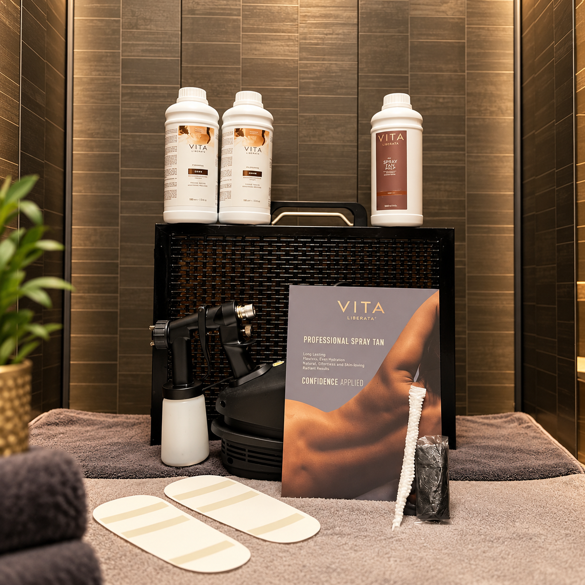 Vita Liberate Professional Spar Tan set arranged on a spa towel against a dark wood tiled wall, including three white bottles, a spray tan pump device, a printed brochure, a brush, and two applicator pads.