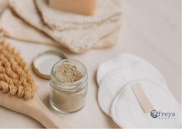 Natural skincare set featuring a wooden body brush with tan bristles, a small glass jar filled with beige powder, reusable cotton pads, a folded beige cloth, and a bar of soap on a light neutral surface.